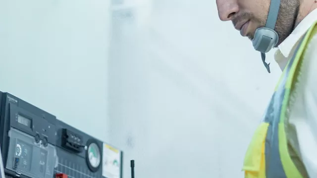 A man wearing a safety helmet and high-visibility waistcoat is standing at an electrical installation with a laptop.