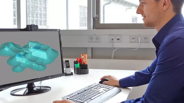 Side view of a man sitting at the desk in front of a PC. The screen shows a 3D visualisation of a building and the representation of the protected volumes.